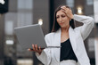 © MZaitsev - A worried businesswoman in a white suit stands outdoors near an office building, holding a laptop and touching her head while dealing with a work problem.