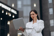 © MZaitsev - A worried businesswoman in a white suit stands outdoors near an office building, holding a laptop and touching her face while dealing with a work problem.