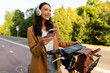 © Home-stock - Happy European woman standing next to electric bike, wearing headphones and holding cellphone while enjoying time outdoors in park