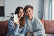 © Daniel - Happy couple at home sitting on sofa holding phone and showing to the camera white blank screen. Copy space on smartphone concept, contracting insurance, internet shops