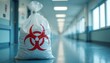 © Viktor - Biohazard waste bag with red symbol rests on hospital floor. Medical rubbish disposal process shown in sterile hallway. Safety and hygiene protocols observed.