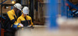 © eakgrungenerd - Two warehouse workers wearing safety helmets reviewing inventory checklist and logistics data in distribution center. Employees inspecting cargo and managing operations.