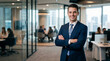© Kaluya Stock - Confident smiling young businessman in a dark blue suit with arms crossed, standing in a modern office with blurred colleagues
