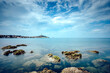 © Travel 'n' Lifestyle - View of mossy rocks emerge from the tranquil turquoise sea towards the distant town under a vast, airy sky, Rovinj, Istria County, Croatia.