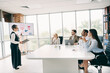 © deagreez - Businesswoman Addresses Team During Presentation in Modern Office with Large Windows and Technology