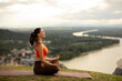 © Home-stock - Side view of young woman sitting cross-legged on mat, focusing on her breath and practicing yoga outdoors, with river flowing on background