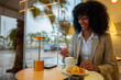 © DusanJelicic - Woman with dark curly hair stirs coffee and enjoys a fresh croissant by a cafe window, smiling and relaxed in business casual attire with an urban morning backdrop