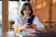 © Pichsakul - Asian young adult woman smiles engaging smartphone cozy cafe table headphones online communication digital browsing technology modern lifestyle leisure education