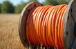 © Viktor - Large orange cable spool rests in dry grass field. Wooden reel holds coiled wire for infrastructure connection. Outdoor rural setting implies telecom or power grid work.