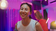 © tirachard - Diverse multi ethnic young adult Asian lady wear futuristic neon glowing glasses smile and drink alcohol beer fun enjoy hangout party in living room night indoor home. Disco lights illuminating house.
