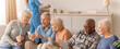 © Prostock-studio - A group of older adults sits together in a living room. They share smiles and laughter while looking at a smartphone. A caregiver is in the background attending to other tasks.