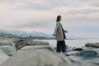 © SHOTPRIME STUDIO - A woman in a long coat stands on rocky shore facing the calm sea, mountains in the distance under cloudy sky. Concept of solitude, nature, travel, and peaceful contemplation outdoors.