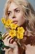 © VICHIZH - Young woman with long blonde hair holding yellow flowers, expressing serenity and beauty against a soft blue background, ideal for seasonal themes.