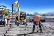 © ramani - Construction workers preparing a concrete surface at a building site. One worker uses a power tool while wearing an orange helmet, with an excavator, cement mixer truck, and tools nearby. 🚧🏗️