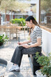 © bongkarn - Asian woman holding pen writing in notebook and sitting cross legged on wooden bench in garden cafe.