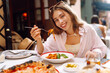 © maxbelchenko - A happy woman eats fresh Italian pasta at a cafe table. A young tourist enjoys spaghetti on a sunny day on the terrace. Concept of food, enjoyment.