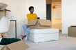 © wavebreak3 - African American woman wearing yellow top sitting on white ottoman handling moving box with phone