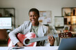 © AnnaStills - Portrait of young adult Black woman smiling while playing acoustic guitar at desk in modern office setting, showing joyful expression and engaging with musical instrument indoors