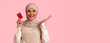 © Prostock-studio - A woman is in a studio holding a red card in one hand and gesturing with the other hand. She is smiling and wearing a hijab. The background is plain pink.