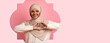 © Prostock-studio - A woman is smiling while wearing a hijab. She is making a heart shape with her hands. The background is pink and simple. Her expression shows happiness and warmth towards viewers.