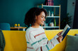 © deagreez - A woman reads a book on a bright yellow couch at home in a cozy living room with modern shelves and colorful decor