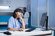 © DC Studio - Fatigued female practitioner in modern hospital office, using digital technology for collaboration and communication. Dedicated nurse in blue scrubs tiredly reviewing medical data on desktop computer.