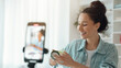© Prostock-studio - A woman smiles while applying a skincare product in a well-lit room. A camera is positioned in front of her, capturing the moment. She seems focused and engaged with her activity.