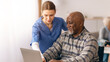 © Prostock-studio - A young woman in blue scrubs teaches an older man how to use a laptop in a care facility. They are both engaged in the activity. Other residents are visible in the background.
