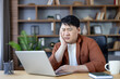 © Liubomir - Asian man looking frustrated and overwhelmed while working on a laptop at his home office desk, experiencing stress, confusion, and difficulty with a work problem
