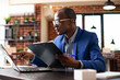 © DC Studio - Black male manager seated at desk concentrating on business project plan, using laptop and company documents for assistance. Businessman holding clipboard and looking at office information on device.