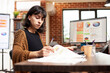 © DC Studio - Young businesswoman sitting in bright brick wall room, reviewing startup company documents on desk. Computer screen displays financial charts as female manager researching business project ideas.