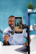 © DC Studio - Smiling blogger on smartphone screen live streaming on social media channel. Young cheerful african american man creating digital content while recording video on mobile phone