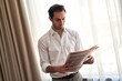 © Teodor Lazarev - Professional man reading morning newspaper, engaging with daily news and current events, seeking information while standing indoors with curtains, portraying a well-informed lifestyle