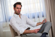 © Teodor Lazarev - Man in a white shirt and plaid trousers holding a newspaper while sitting on a chair, projecting a serious and confident expression in a modern interior