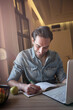 © Teodor Lazarev - Man smiling, writing notes in a notebook while working on a laptop at home, focusing on productivity, inspiration, and distance learning concepts