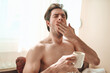 © Teodor Lazarev - Man feeling exhausted and sleepy in bed, yawning and covering his mouth while holding a warm coffee cup, experiencing morning fatigue and preparing for a new day