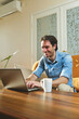 © Teodor Lazarev - Man enjoying flexible remote work, typing on a laptop with a coffee mug at home, comfortably managing his business in a modern living space