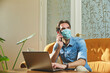 © Teodor Lazarev - Man wearing a protective medical mask, sitting on a sofa while talking on a smartphone and using a laptop, performing remote work indoors during social distancing