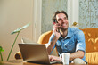 © Teodor Lazarev - Young man happily working from home, talking on smartphone, smiling while looking up, connecting with comfort and technology from his residential office couch