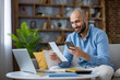 © Liubomir - Young man smiling in a cozy home office, holding a bill and smartphone while managing online banking, digital payments and budgeting with modern tech and laptop nearby