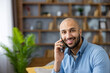 © Liubomir - Young bald man engaging in a happy phone conversation, smiling brightly while at home, showcasing modern communication and connection in a casual setting