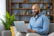 © Liubomir - Young bald man smiling, using a laptop while sitting on a comfortable couch in a living room, happily engaging in remote work and managing online tasks