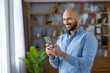 © Liubomir - Smiling bearded bald man in a blue shirt using his smartphone indoors, happily texting and browsing, illustrating modern digital communication, connectivity and casual work-life lifestyle
