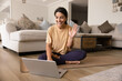 © fizkes - Cheerful Latina woman sitting cross-legged on yoga mat in front of laptop, smiling and waving at screen during online yoga or meditation practice from home. Modern tech usage, virtual workouts session