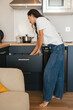 © sutulastock - Young woman in casual jeans and white T-shirt bending over an open kitchen drawer, searching for utensils in a modern home interior during a relaxed morning routine with natural light.