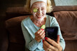 © Geber86 - Smiling senior woman doing home spa facial while taking a selfie on the sofa at home