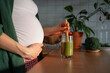 © wifesun - Pregnant woman gently holding her baby bump while stirring a fresh green smoothie in the kitchen, focusing on healthy eating and vital nutrition for maternal well-being