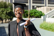 © wavebreak3 - African American woman holding rolled diploma, smiling on campus plaza with cap, gown, watch