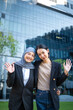 © BJ Day Stock - Portrait of two smiling diverse Asian businesswoman, one wearing hijab, happily waving hands to the camera outdoors against a modern office building and green trees background. Vertical Shot
