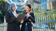 © BJ Day Stock - Portrait of two smiling diverse Asian businesswoman, one wearing hijab, discussing work on a tablet together outdoors against a modern office building and green trees background.